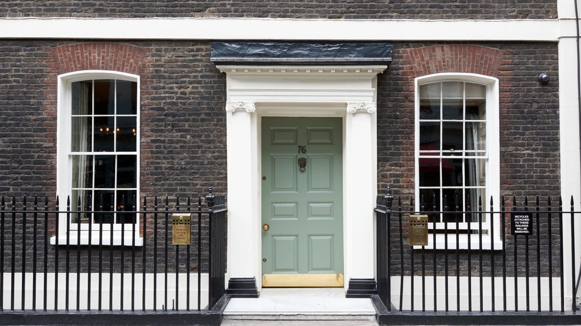 A mid-shot of a green door, within two black gates to the left and right. The door is positioned in the centre of the house, which is made from dark brown brick with two white bay windows.