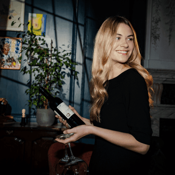 Image of a blonde waitress carrying a bottle of wine and two wine glasses in a restaurant at Dean Street. The room is dimly lit with a shadow from the window cast in the background.