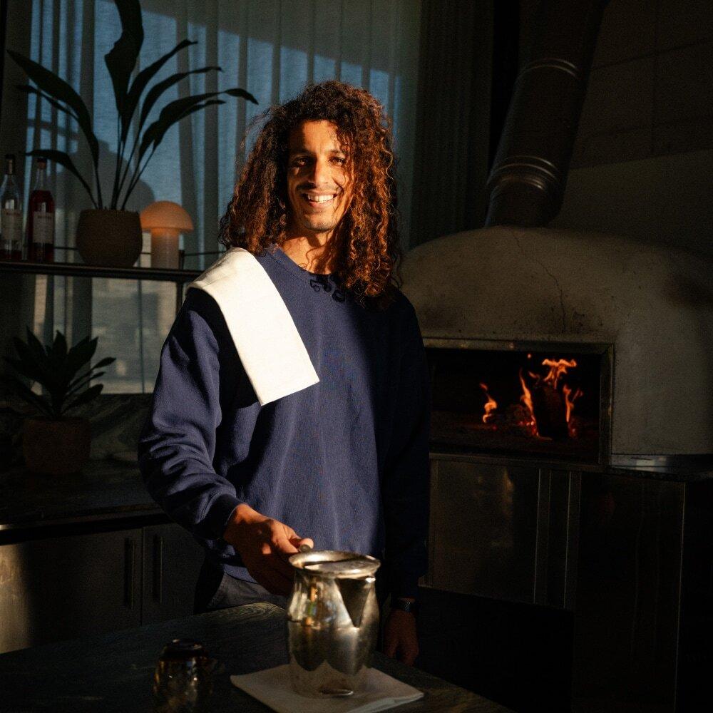Mid shot of a man smiling in a sunlit room. The man is wearing a blue jumper and has a white serving cloth positioned over his right shoulder. He is holding a silver water jug.
