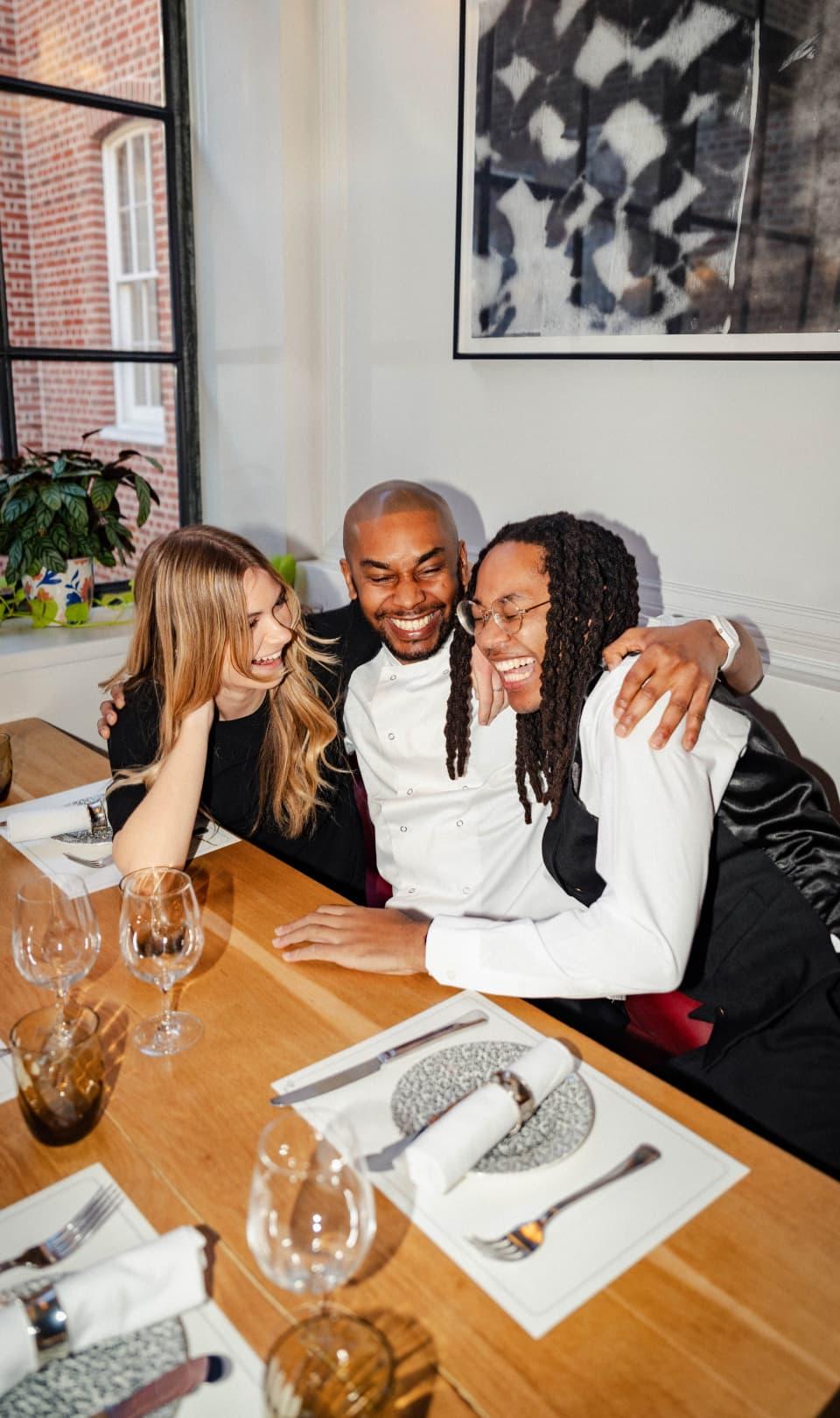 Image of 3 people sitting at a table with glasses laughing. The group is multicultural and wears a combination of white and black clothing. The background contains a window on the left and a white wall on the right with a black-and-white framed image.