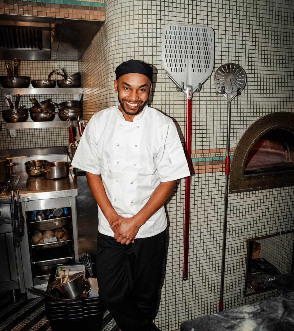 Image of a male chef in chef whites standing in the centre of the frame with his hands and legs crossed. In the background, there is the pizza oven and the kitchen equipment from Cecconi's pizza bar.