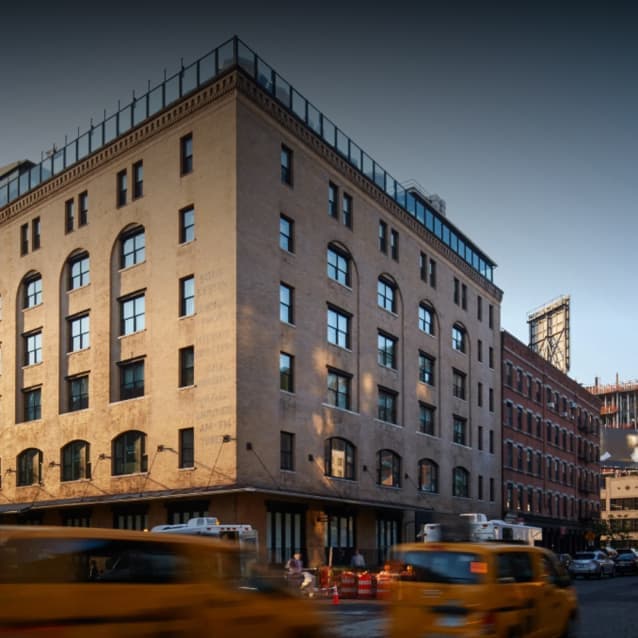 Corner view of a historic multi-story brick building with arched windows, rooftop railing, and yellow taxis passing by, set against a cityscape with a bridge in the background