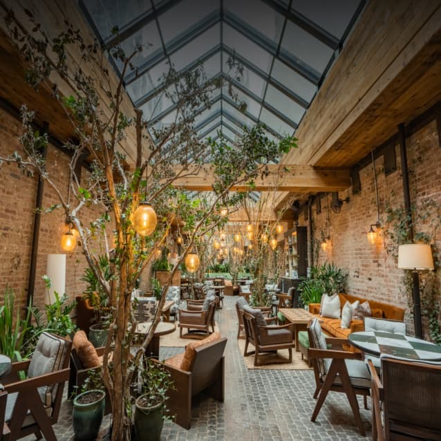 Spacious indoor restaurant with a greenhouse-style glass ceiling, exposed brick walls, wooden beams, hanging amber lights, lush potted plants, and cozy wooden seating arrangements