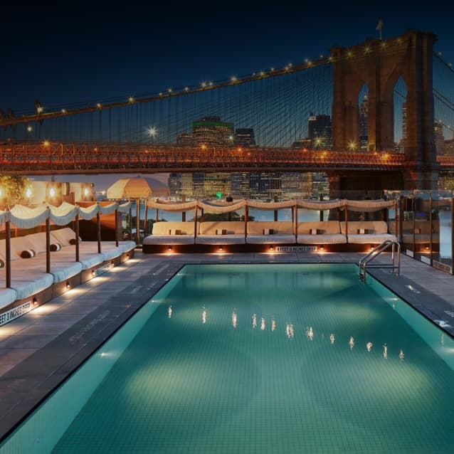 Rooftop pool at night with plush poolside beds, string lights, and the illuminated Brooklyn Bridge in the background, overlooking the New York City skyline