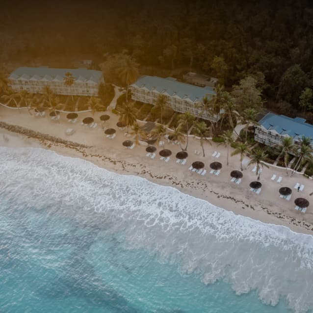 Aerial view of a beachfront resort with pale blue buildings, palm trees, thatched umbrellas, and lounge chairs, set against turquoise waves and dense tropical greenery