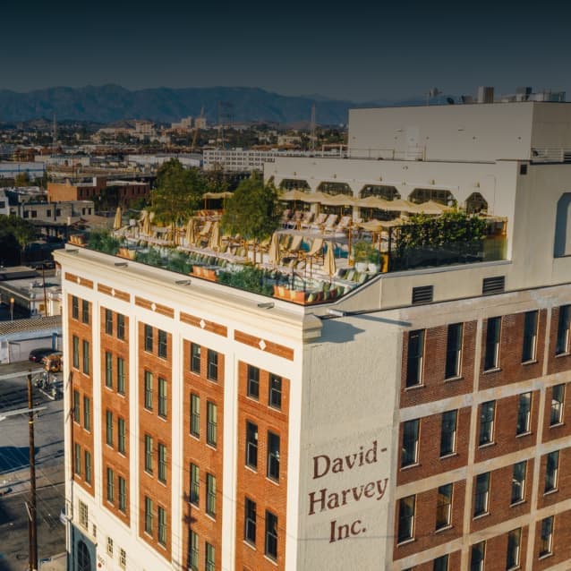Brick and white building with "David-Harvey Inc." painted on the side, featuring a rooftop pool and lounge area with greenery, against a backdrop of mountains and cityscape