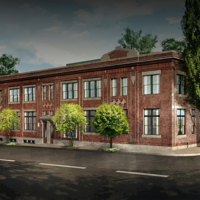 Two-story red brick building with large rectangular windows, decorative brick patterns, and small trees lining the sidewalk