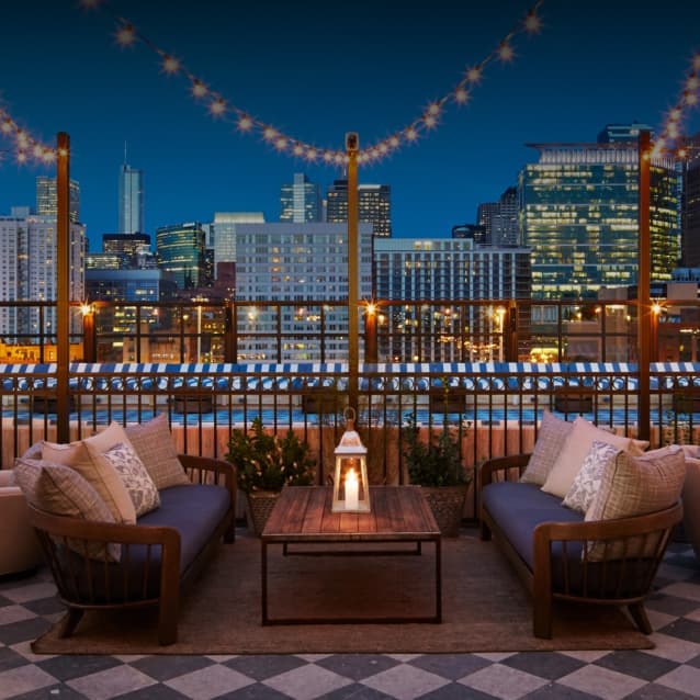 Rooftop lounge with string lights, cozy seating, and a lantern on a wooden table, overlooking a city skyline at night