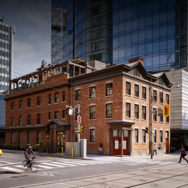 Historic red-brick building with a rooftop terrace, surrounded by modern glass skyscrapers at a city intersection