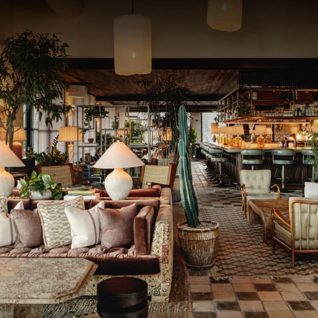 Lush lounge with eclectic seating, indoor plants, patterned cushions, large ceramic lamps, and a bar area in the background