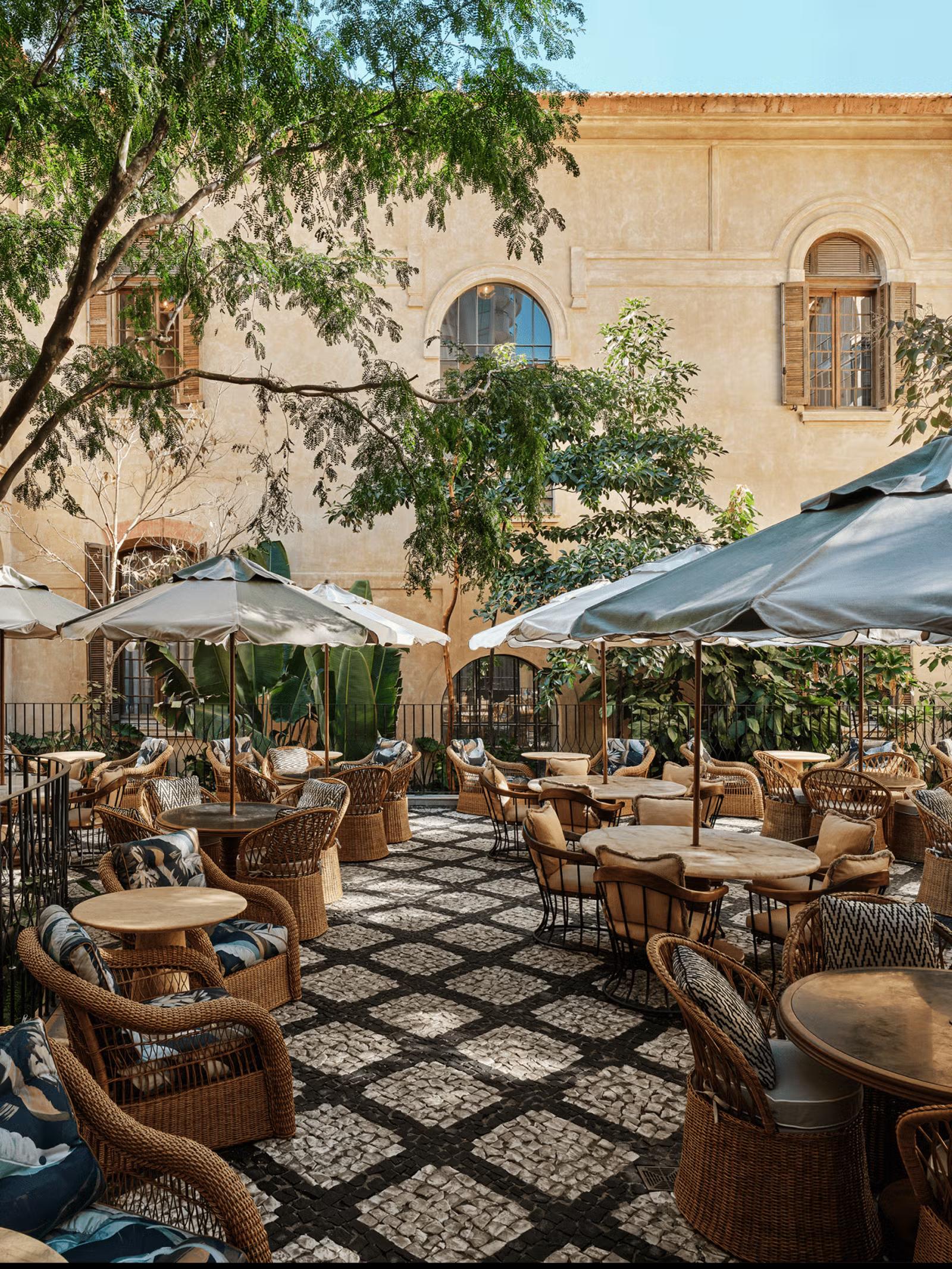 Outdoor table and chairs with umbrellas and cushions in a patio courtyard. In the background there isa architectural building with trees framing the courtyard